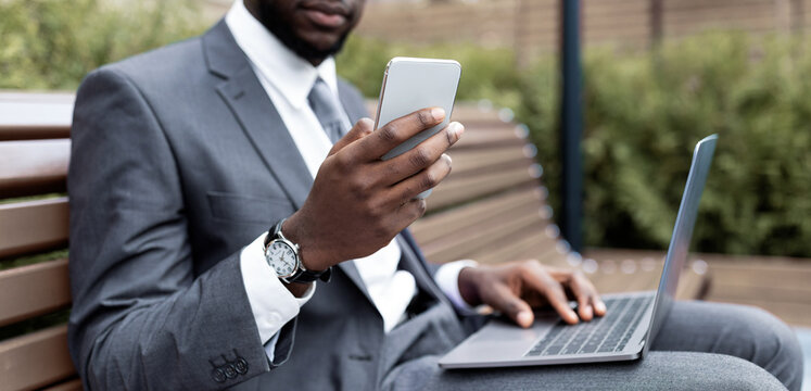 A business professional sits on a park bench, focused on both a laptop and a smartphone. Wearing a suit, he is engaged in work while enjoying the outdoor atmosphere.
