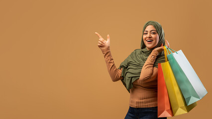 An excited Muslim woman is holding colorful shopping bags and pointing to an empty space with a smile. She showcases a welcoming attitude, perfect for a shopping advertisement.