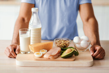 Fresh ingredients for a nutritious meal are arranged on a wooden cutting board. A person prepares eggs, chicken, milk, cheese, avocado, spinach, and nuts for a balanced diet.