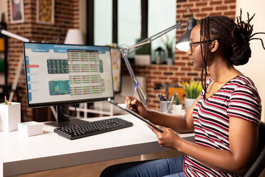 Self employed woman analyzing business charts on desktop pc, comparing metrics with notes from clipboard. Black female manager reviewing financial graphs on computer screen at home. - Powered by Adobe