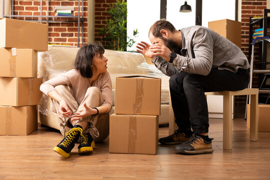 Husband and wife sit surrounded by cardboard boxes in brick wall apartment, having heated argument over arranging items. Tense moment between white couple during stressful moving day at new home. - Powered by Adobe