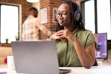 Self employed woman seated with headphones, attentively listening to online webinar on laptop....