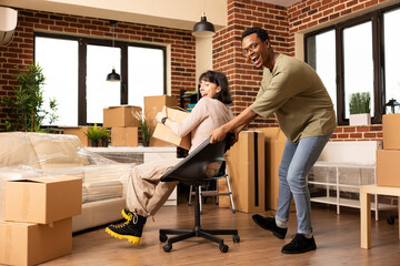 Cheerful young couple celebrating moving day as woman holding box is pushed on chair by african american husband in new apartment. Excited diverse married individuals having fun in unfurnished room.