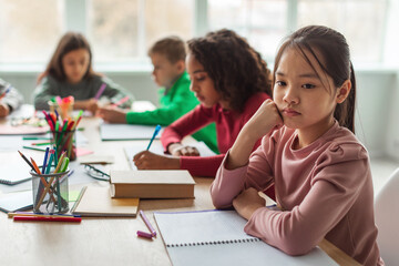 In a modern classroom, an unhappy Asian schoolgirl is deep in thought, feeling troubled by education challenges, while her diverse classmates are focused on their learning tasks at their desks.