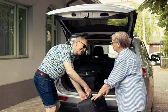 Old smiling man and woman packing their luggage into the vehicle trunk, both retired and excited to leave on a summer getaway. Happy couple reflecting the joy of travel, retirement activity.