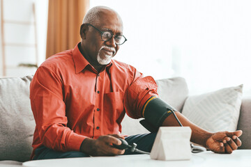 An older gentleman wearing glasses measures his blood pressure at home. He sits comfortably on a couch, focusing on the monitor. Natural light fills the room, creating a peaceful atmosphere.