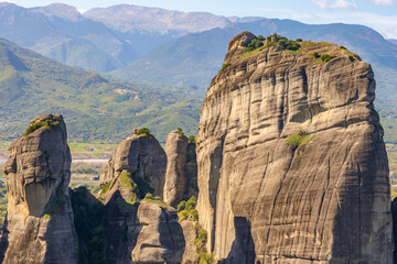 Close-Up View of the Towering Sandstone Pillars of Meteora, Greece, Featuring a Prominent Vertical Rock Face and a Distant Mountainous Background under a Blue Sky