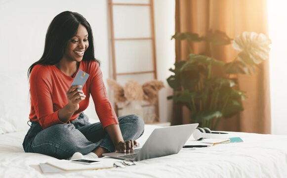 A woman sits comfortably on her bed, smiling as she holds a credit card while using her laptop. The cozy bedroom features warm lighting, plants, and various items scattered around. - Powered by Adobe