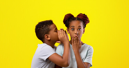 Two children are whispering together against a vivid yellow background. One child leans in closely as the other listens with a surprised expression, creating a playful moment.