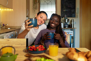 Happy young multiracial women smiling, holding coffee mugs, and having a healthy breakfast in their kitchen