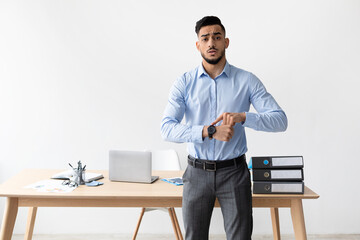 A serious Middle Eastern male manager stands in a bright office, pointing at his wrist watch. He looks directly at the camera, communicating urgency and reminding others about deadlines.