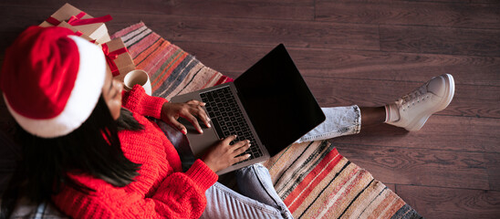 A young woman wearing a red festive sweater sits on a patterned rug, using a laptop. She is surrounded by holiday gifts and enjoys a warm drink. The setting is cozy and inviting.