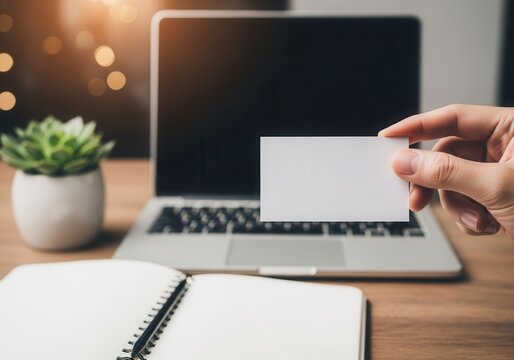Hand holds blank business card in front of laptop and notebook