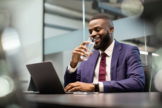 Positive young black man manager drinking water while working on laptop, typing on computer keyboard, making marketing research or sending emails, office interior, copy space