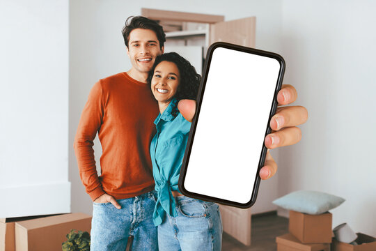Two smiling individuals stand in a cozy, uncluttered room, showcasing a smartphone with a blank screen. They appear joyful and content in their new living space. - Powered by Adobe