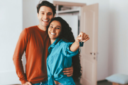 A joyful couple stands close together, smiling as they hold up keys in a well-lit, modern room, celebrating their new home and looking forward to their future together.