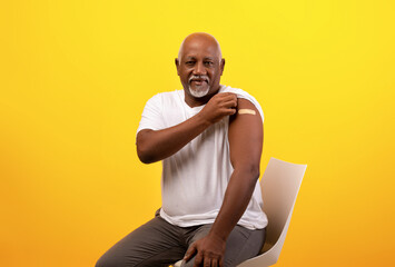 A positive elderly black man shows a bandage on his shoulder after receiving the Covid-19 vaccine. He is seated against an orange background, smiling at the camera, displaying his vaccination pride.