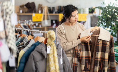 Latina woman looking at outerwear, jackets and coats at a sale in a clothing store, she pays attention to the price tag. Against the background are hangers with jackets and signs about discounts. © JackF