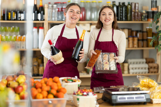 Young smiling saleswomen offer to buy wine and champagne for the holiday. Supermarket workers are holding new arrivals of sweets and wine in their hands.