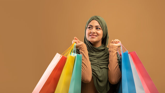 A smiling muslim woman in a hijab stands against a brown background, holding colorful shopping bags while looking aside. She embodies joy and excitement for seasonal sales and discounts.