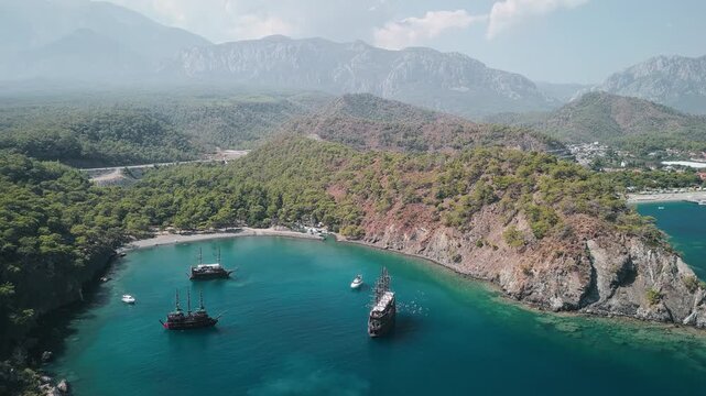 Aerial view of Phaselis Bay with sailing ships and Mount Olympos in the background. Clear blue water and scenic Mediterranean coastline near Antalya, Turkey. Captures scenic coastal beauty in Turkey.