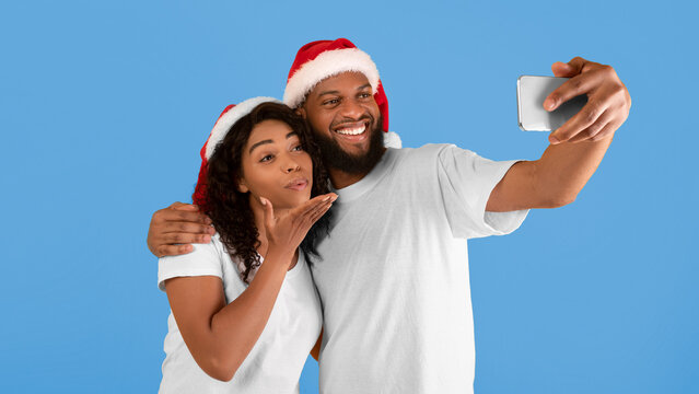 Playful african american spouses in Santa hats taking selfie on smartphone, woman blowing air kiss to cellphone camera, posing on yellow studio background - Powered by Adobe