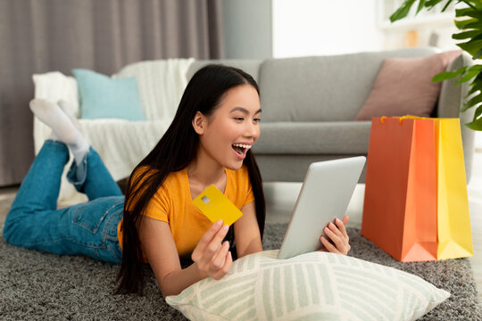 Overjoyed young asian lady lies on the floor surrounded by shopping bags, excitedly using a tablet and credit card to shop online. Her delighted reaction shows her enjoyment of the sale.