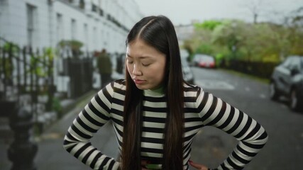 Woman standing with arms akimbo on an urban street shows discomfort, while wearing a striped sweater outdoors in a city setting with a blurred background and expressive face.