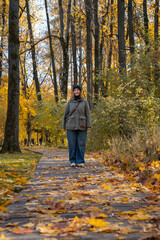 Fototapeta premium Woman walks through an autumn park in warm sunlight, wearing a khaki jacket and jeans surrounded by yellow leaves