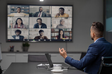 An Arabic CEO conducts an online meeting with his multiracial team, discussing strategies while seated at a table. The team engages remotely via a large TV display in a contemporary office space.