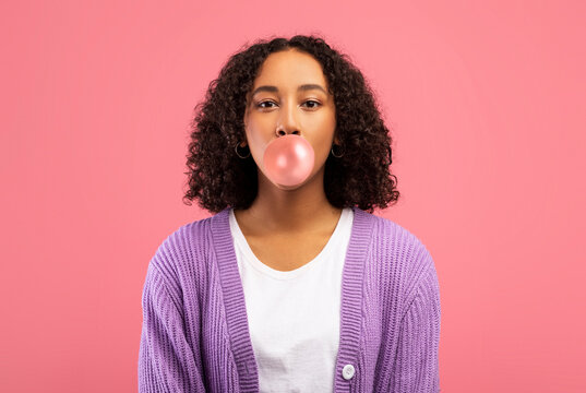 A pretty young black woman is blowing a bubble with her gum while looking directly at the camera. She is casual in her style, set against a vibrant pink studio background.