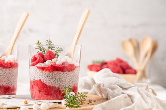 Two glasses of layered raspberry and chia seed pudding topped with fresh raspberries and white crowberry on a rustic wooden platter