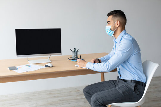 Young Middle Eastern man in a surgical face mask sits at a desk, focused on a laptop with a blank screen. Papers are scattered nearby, indicating remote work and study activities in an office setting.