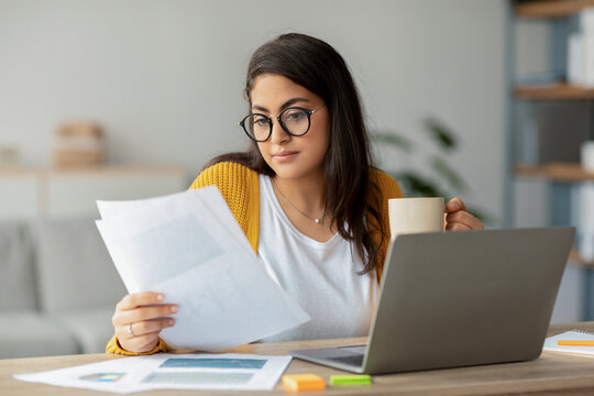 Concentrated arab businesswoman sits at her home office desk, reading papers while holding a cup of hot beverage. She is engaged in her work in a cozy living room setting.