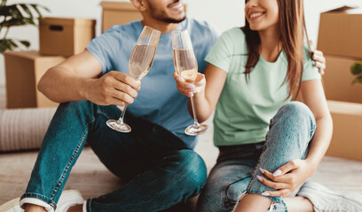 A joyful couple sits on the floor, holding glasses of sparkling drink and smiling at each other. They are surrounded by moving boxes in their new home, celebrating together.