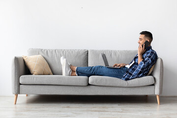 A smiling Arab man sits comfortably on a sofa at home, engaging in a phone call while working on his laptop. He enjoys his distance job, showcasing a positive attitude and relaxed environment.