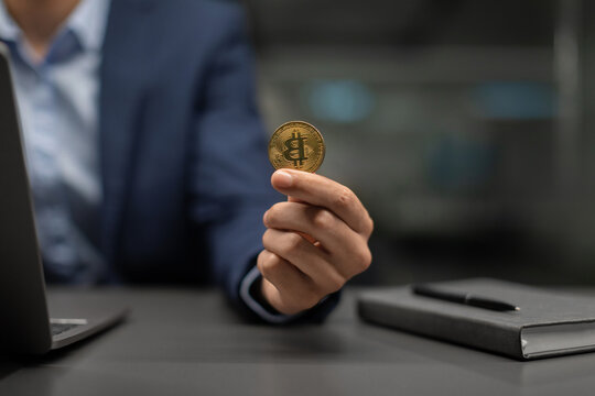 Business professional dressed in a suit holds a bitcoin while using a laptop at an office desk. The scene emphasizes investment and trading activities in a contemporary environment.