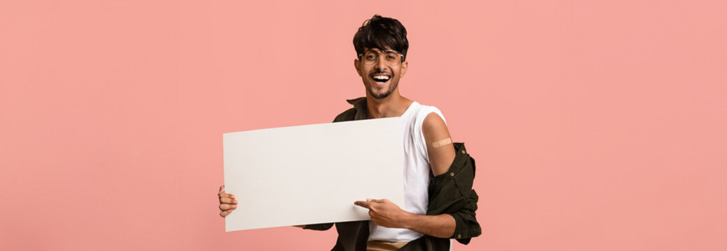 A young man stands against a pink backdrop holding a large blank sign. He is smiling and wearing a sleeveless shirt with a casual jacket draped over his shoulders, showcasing a friendly demeanor.