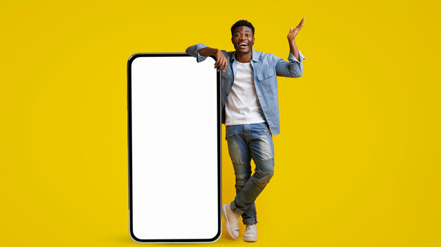 A cheerful young man stands next to a large blank smartphone against a vivid yellow backdrop. He is smiling and gesturing with excitement, showcasing joy and enthusiasm. - Powered by Adobe