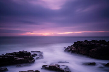 Purple dusk sky over rocky shoreline with misty waves from long exposure, dramatic coastal landscape background suited for travel, wellness ads.