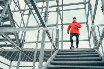 Smiling runner man dressed bright red softshell sporty clothes running down by huge steel industrial stairs in cold winter day. People healthy lifestyle concept photo.
