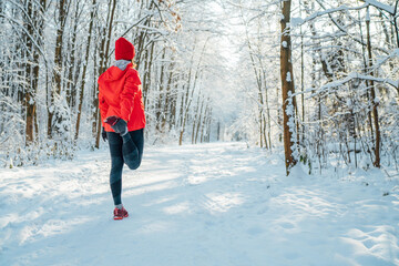 Trail runner woman dressed bright red windproof jacket warming up doing streching in  picturesque snowy forest path during sunny frosty day. Sporty active people and winter training concept image.