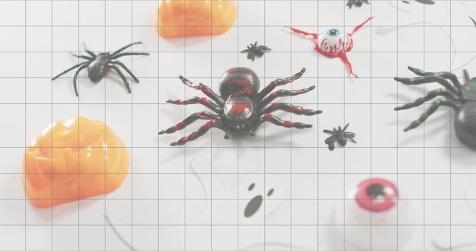 Sitting glossy black-and-red plastic spider on white grid mat at table, with pumpkins and eyeballs