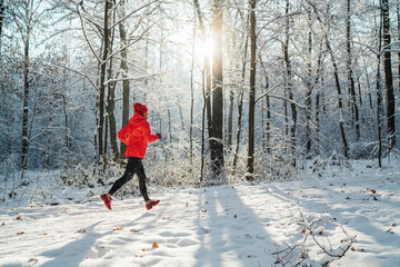 Trail runner woman dressed bright red windproof jacket jogging picturesque snowy forest path during sunny frosty day. Sporty active people and winter training concept image.