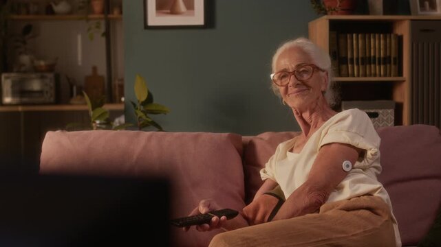 Medium shot of happy elderly Caucasian woman, wearing GCM patch on arm to monitor diabetes, relaxing on couch at home, turning on television set with remote control to watch favorite show