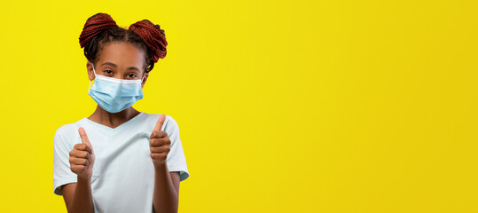 A young girl with braided hair in two buns wears a light blue shirt and a face mask. She is smiling and giving thumbs up against a cheerful yellow background, showing positivity.