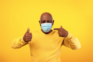 A senior black man wearing a yellow sweater and a face mask points to the mask while giving a thumbs up.