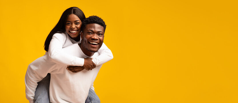 A man gives a woman a piggyback ride, both smiling widely with happiness. They wear casual white tops and have a vibrant yellow background, creating a fun and cheerful atmosphere. - Powered by Adobe