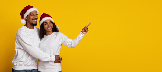 Fototapeta premium Two smiling individuals wear Santa hats and embrace joyfully. They stand against a vibrant yellow background, with one pointing excitedly. The scene captures holiday cheer and warmth.