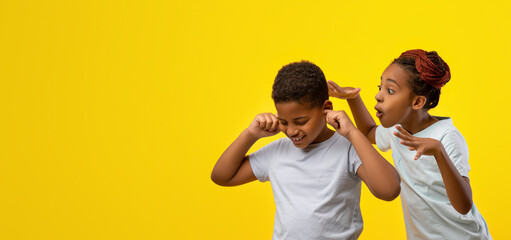 A boy and a girl share a joyful moment in front of a vibrant yellow backdrop. The girl pretends to whisper something funny, causing the boy to react with a playful expression.